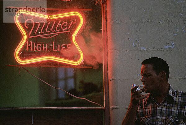 A man lights a cigarette by a neon sign.