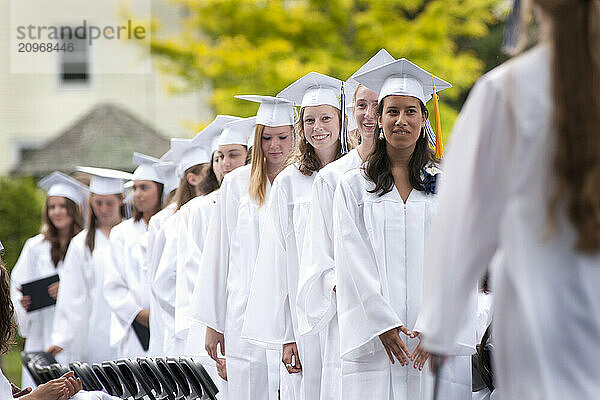 Female students in white caps and gowns line up to receive their diplomas at their high school graduation ceremony.