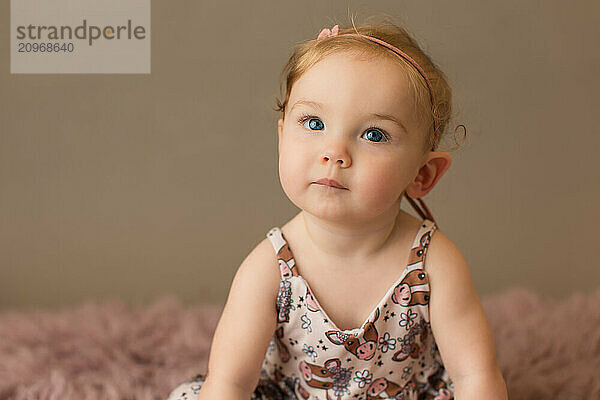 Thoughtful baby with blue eyes in a patterned dress and pink hea