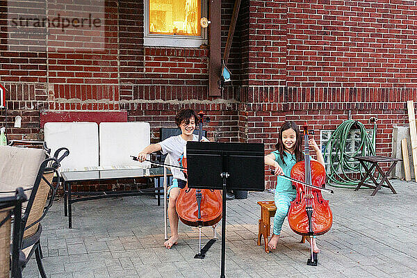 Two smiling children play cello together on home patio