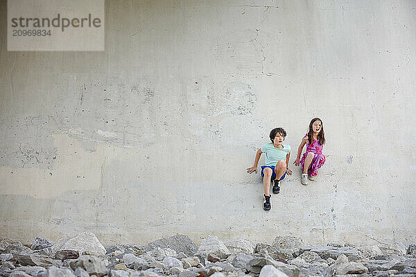 Two children sit against tall cement wall in a barren urban landscape