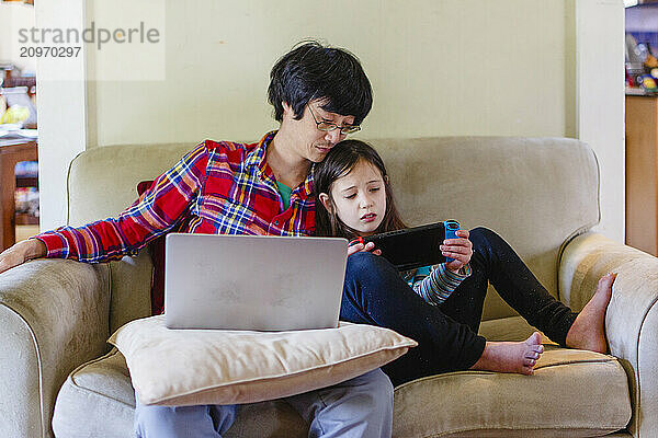 A father sits on couch next to small child working on computer