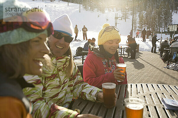 Friends in ski gear socialize and enjoy a beer on the deck on a sunny day on the mountain.