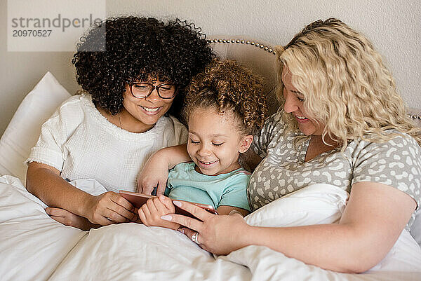 Mother and daughters cuddle in bed smiling looking at a tablet