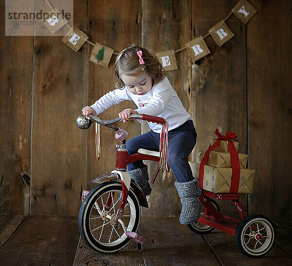 Toddler carrying gifts on red tricycle Christmas backdrop