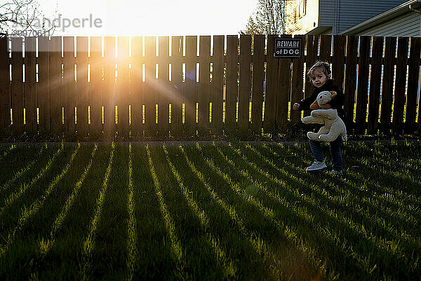 Young girl with stuffed dog walking past sunny fence