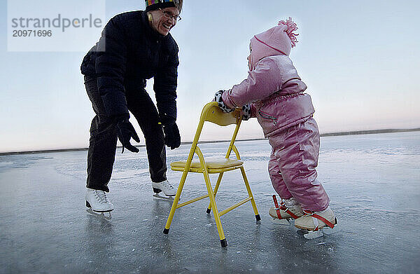 A mother is filled with joy as her daughter is learning to ice skate as she balances with the aid of a chair.