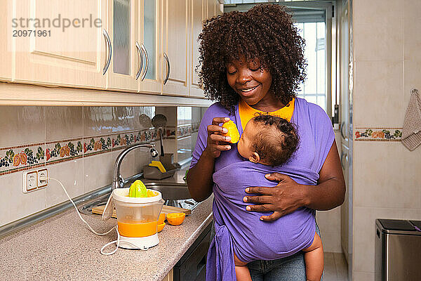 Mother holding her baby is making orange juice in the kitchen