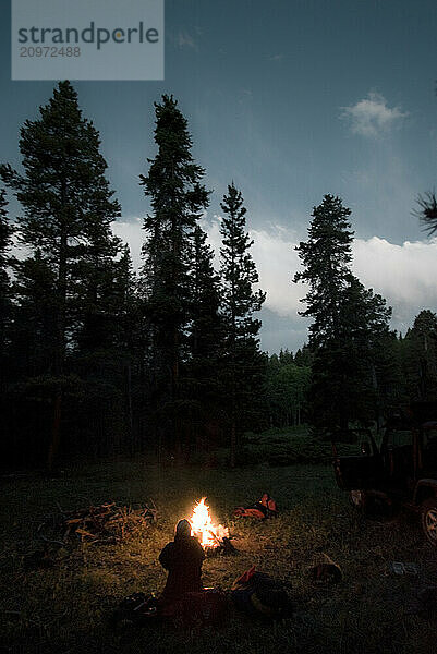 Silhouette of a young woman sitting next to a camp fire at dusk.