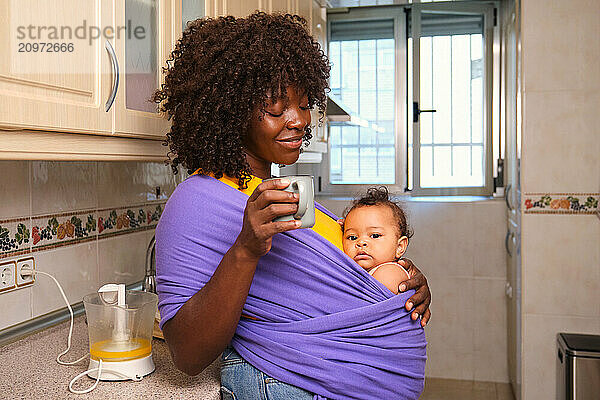 Young mother holding her baby is enjoying a cup of coffee in kitchen