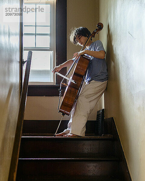 A tween boy stands in staircase playing cello