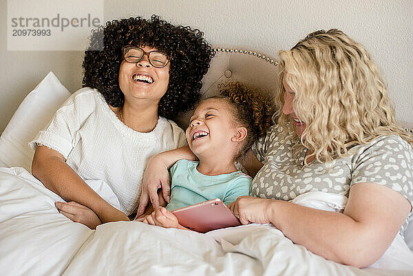 Mother and daughters are laughing while holding a tablet in bed