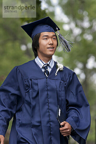 A young male student walks confidently in cap and gown after having received his diploma at his high school graduation ceremony.