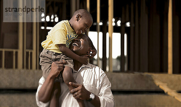 A son sits on his father's shoulders and laughs while they watch their new home constructed.