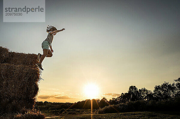 Young girl jumping from straw bales at sunset