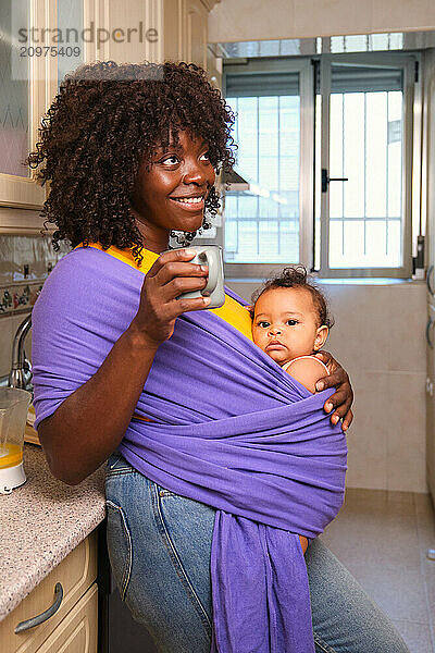 Mother holding her baby in a wrap enjoying a cup of coffee in kitchen