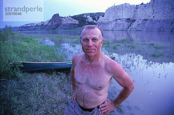 A shirtless man smiling by a river side.