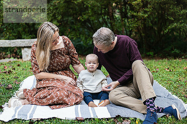 Mom and dad smile down at toddler boy as they sit on blanket in grass.