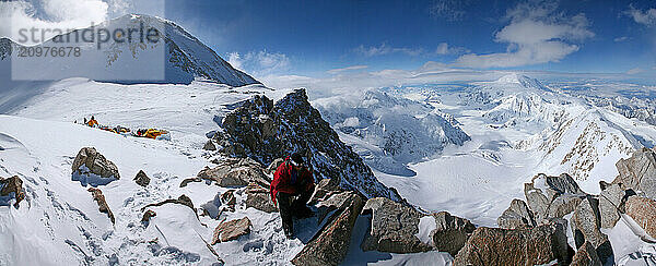 Mountaineer sitting at Denali West Buttress Route High Camp  with summit and 14k camp visible  Denali National Park  Alaska  USA