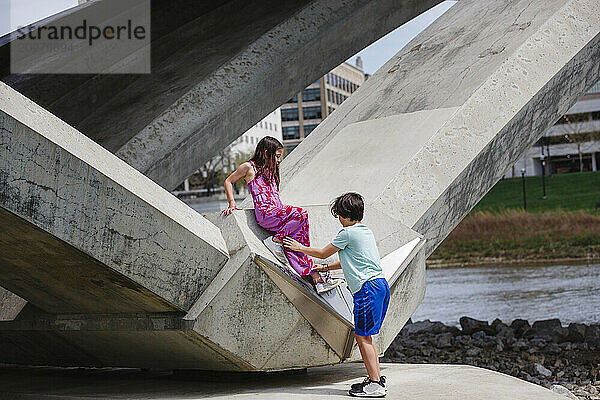 Young boy helps small girl down from bridge piling