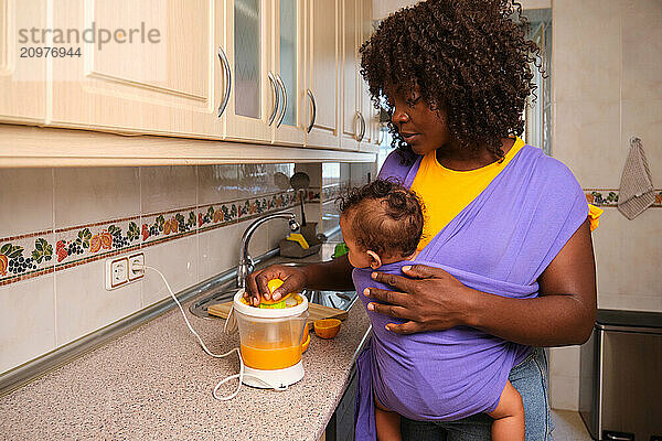 Mother making orange juice carrying her baby in a sling