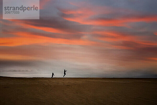 Silhouette young sisters running down sand dune at sunset