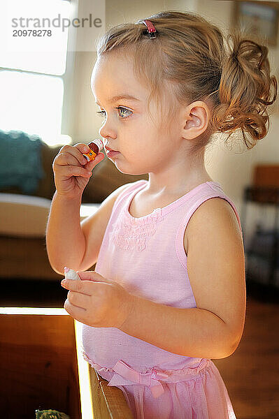 Toddler with curly pigtails putting on lipstick indoors