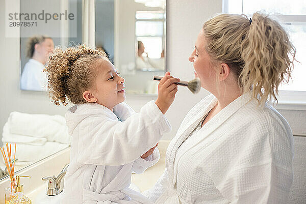 Daughter is brushing make up on mothers face in the bathroom