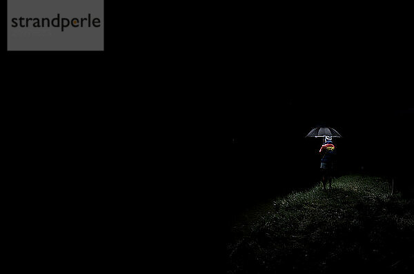 Minimalistic view of a young woman holding an illuminated umbrella on the edge of complete darkness.