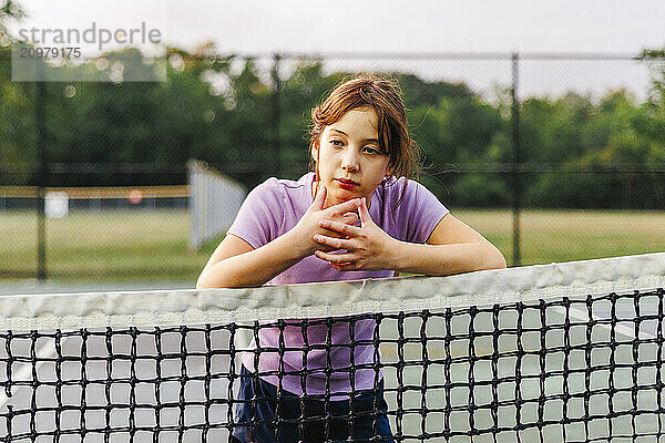 Portrait of thoughtful girl leaning on net at tennis court