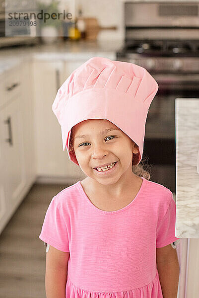 Little girl wearing a pink chef hat and dress in kitchen