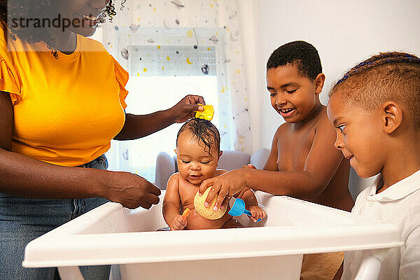 Mother washing baby in small bathtub helped by sons