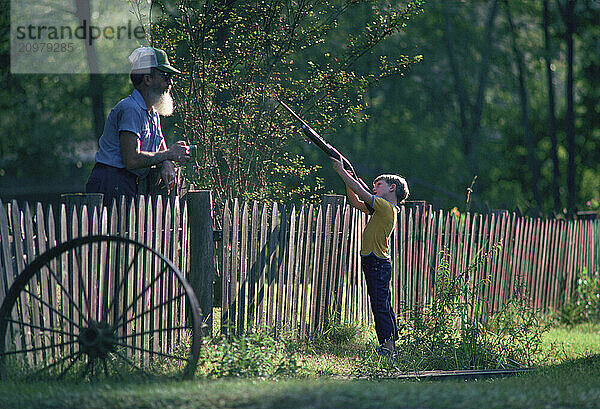 A little boy points a rifle by a picket fence.