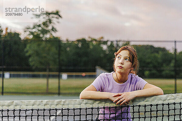 A girl looks leans on net at tennis court at sunset looking up