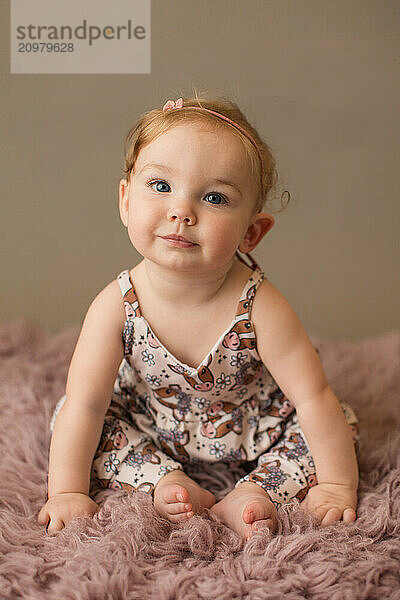 Smiling blue eyed girl in a cow print romper on a pink rug