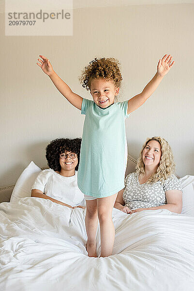 Little girl is jumping on the bed with mother and sister watching her