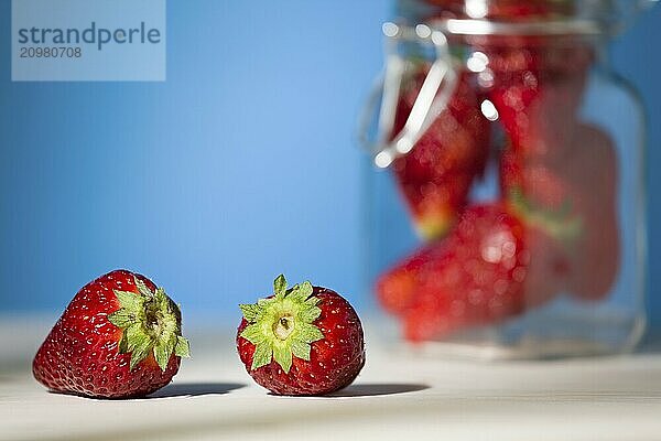 Close up of two strawberries on a table with blue background and a glass jar full of strawberries on the background