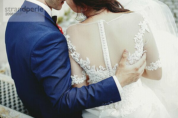 Groom hugs bride back in white dress. Blurred background.