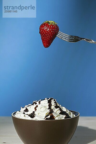 Strawberry suspended from a fork over a cup of whipped cream and chocolate with blue background