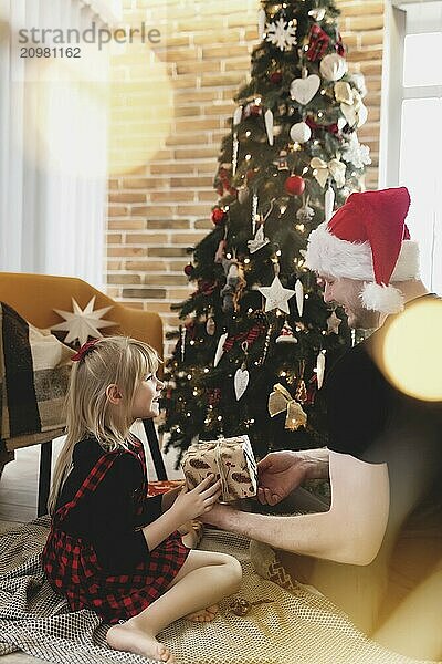 Young father in red Santa hat giving Christmas gift to his smiling daughter near decorated Christmas tree. Girl dressed in festive red-black Christmas outfit. They smiling  happy because of New Year