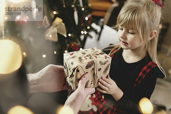 Young father in red Santa hat giving Christmas gift to his smiling daughter near decorated Christmas tree. Girl dressed in festive red-black Christmas outfit. They smiling  happy because of New Year