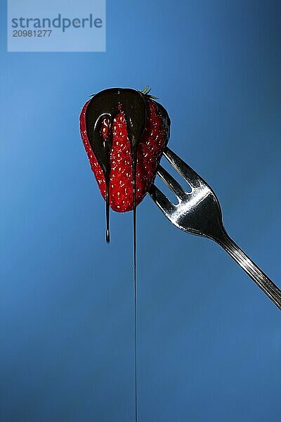 Close up of a strawberry suspended from a fork with liquid chocolate above on a blue background