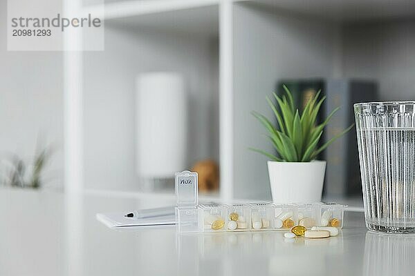 Closeup of medical pill box with doses of tablets for daily take a medicine for treatment  cure the disease. Glass cup of water on table  gray background. Green flowerpot or plant. Pen and notepad