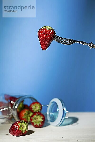 Strawberry suspended from a fork and under a jar of strawberries with blue background