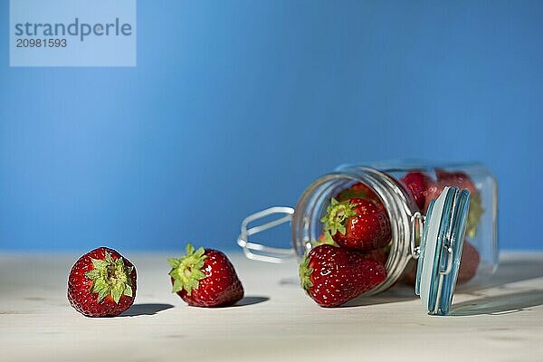 Strawberries and a glass jar full of strawberries lying down on a table with blue background