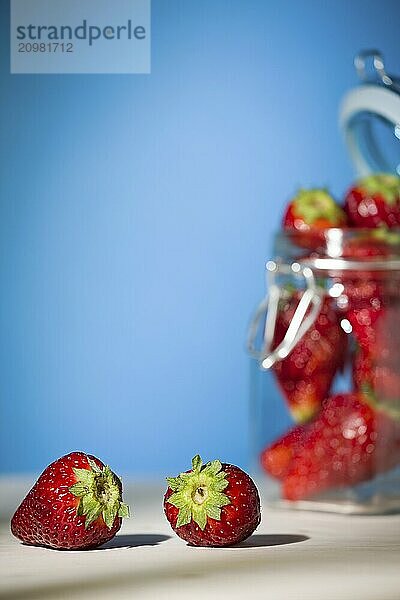 Strawberries on a table with blue background and a glass jar full of strawberries on the background