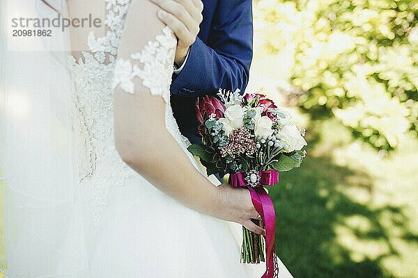 Bride in white dress is holding wedding bouquet. Groom hugs bride. Blurred background.