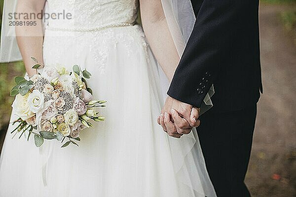 Bride in white dress is holding wedding bouquet. Groom hugs bride. Blurred background.