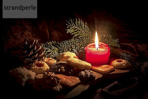 Burning red candle stands between Christmas biscuits decorated on a wooden board against a dark background