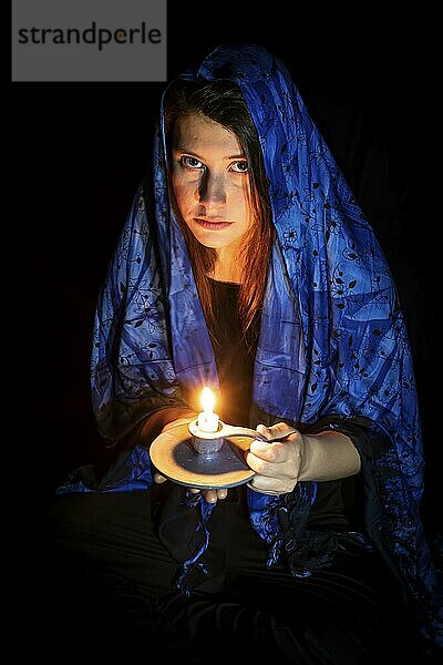 Sad young woman with candle and blue headscarf against a black background